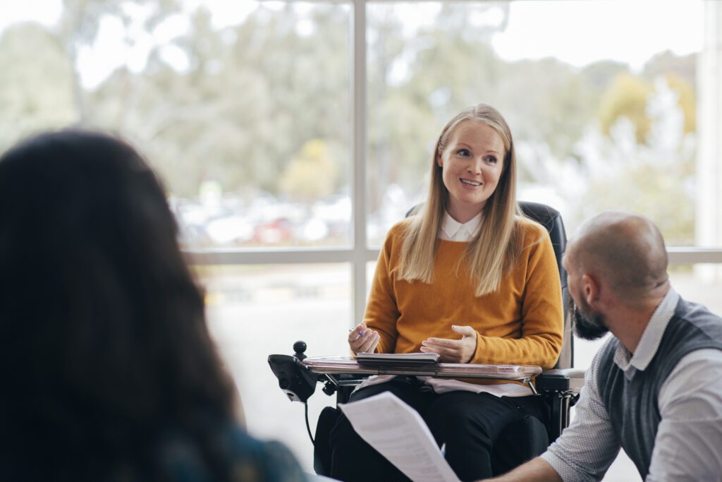 A woman power chair user leading a discussion. She has blonde hair and is wearing a yellow sweater.