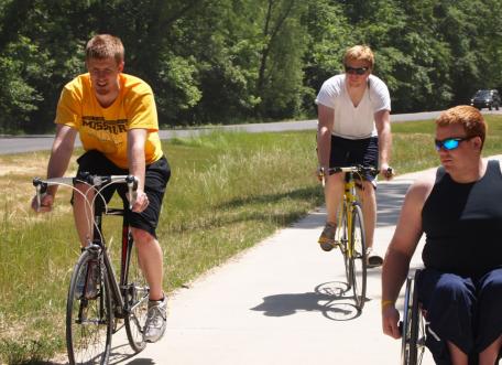 Three young men, two on bikes and one wheelchair user, on a paved trail together.