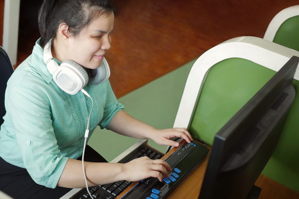 Young Blind Woman With Headphone Using Computer With Refreshable Braille Display. 
